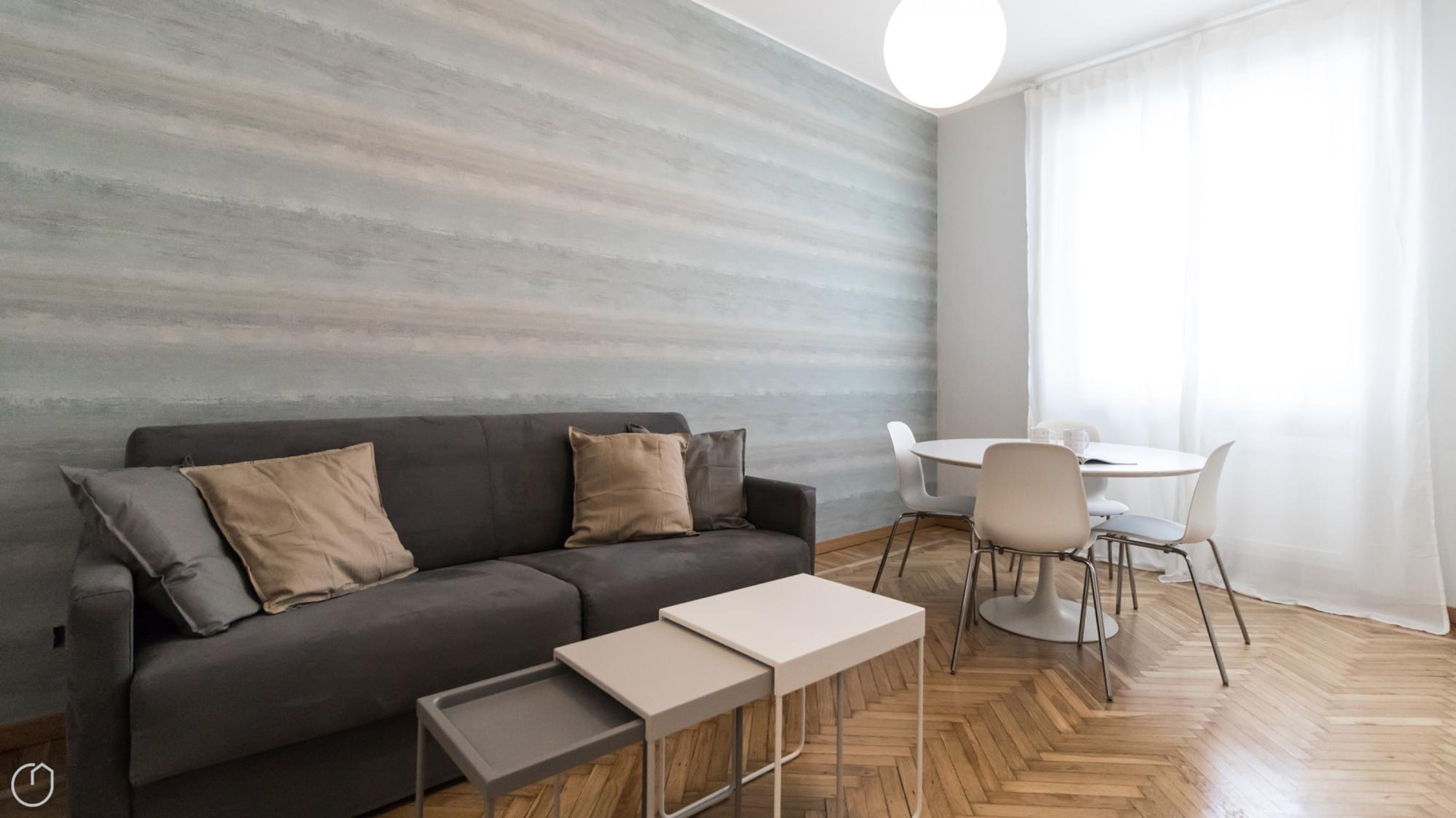 Un salon moderne avec un canapé gris, deux tables d’appoint, une table à manger ronde avec des chaises, et une grande fenêtre ornée de rideaux légers.
