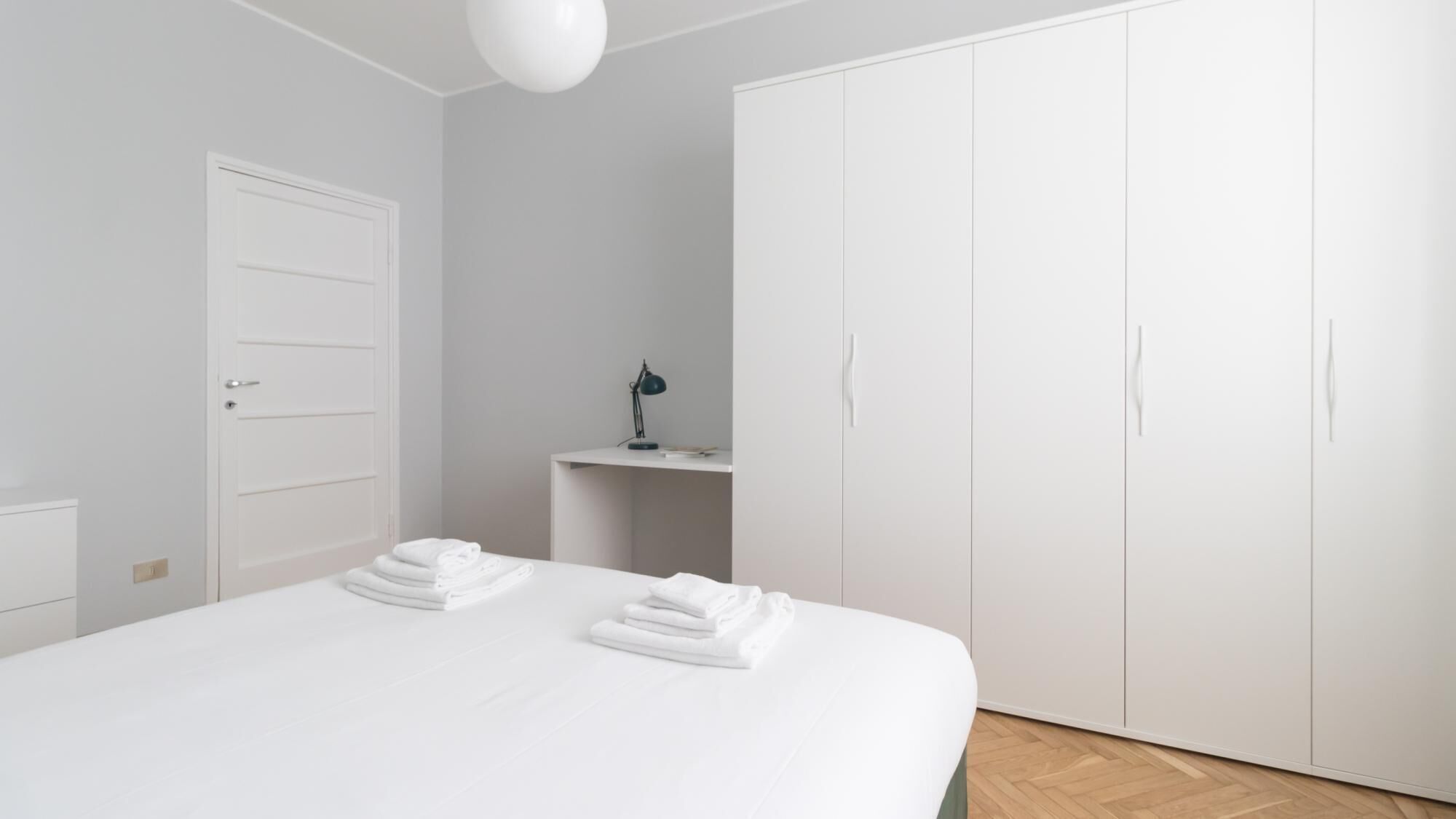 A bedroom with a bed, a wardrobe, a desk with a lamp, and a door.