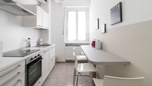 Une cuisine de taille réduite avec des meubles blancs, une cuisinière, un évier et une fenêtre ornée de rideaux légers.