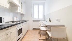 A modern kitchen with white cabinets, a gas stove, a microwave, and a window with blinds. - Italianway Easy - Sacconi 2 A (Milan)