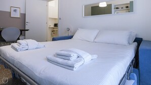 Una cama bien hecha con sábanas blancas y toallas dobladas, una mesita de noche, una silla y un espejo en la pared.
