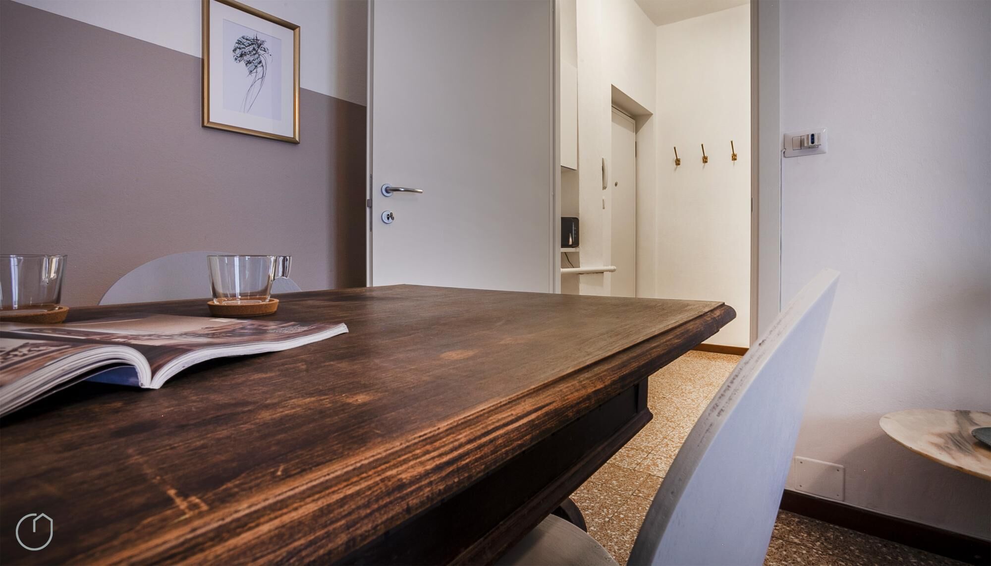 Une table de salle à manger en bois, avec un magazine, deux verres et un dessous de verre. Un tableau encadré est accroché au mur.