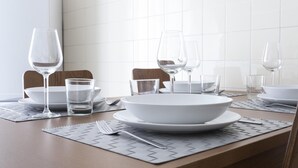 Una mesa de comedor dispuesta con platos blancos, tazones y vasos transparentes, con una computadora portátil a la izquierda.