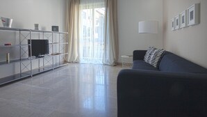 Una sala de estar moderna con un sofá, un televisor sobre una estantería metálica y una ventana con cortinas.