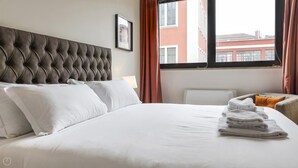 Habitación de hotel con una cama grande, ropa de cama blanca, cabecera capitonada, ventana con cortinas y vistas a edificios.