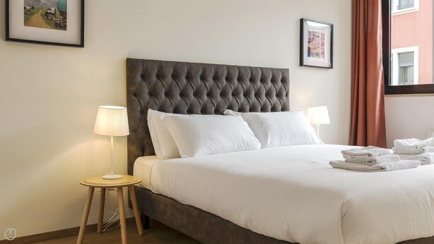 Habitación de hotel con tablero de cabecera acolchado, ropa de cama blanca, dos lámparas de noche y una cama bien hecha con toallas dobladas.