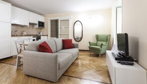 Un espace de vie compact comprenant un canapé gris, un fauteuil vert, un téléviseur à écran plat et un coin cuisine avec des meubles blancs et un four à micro-ondes.