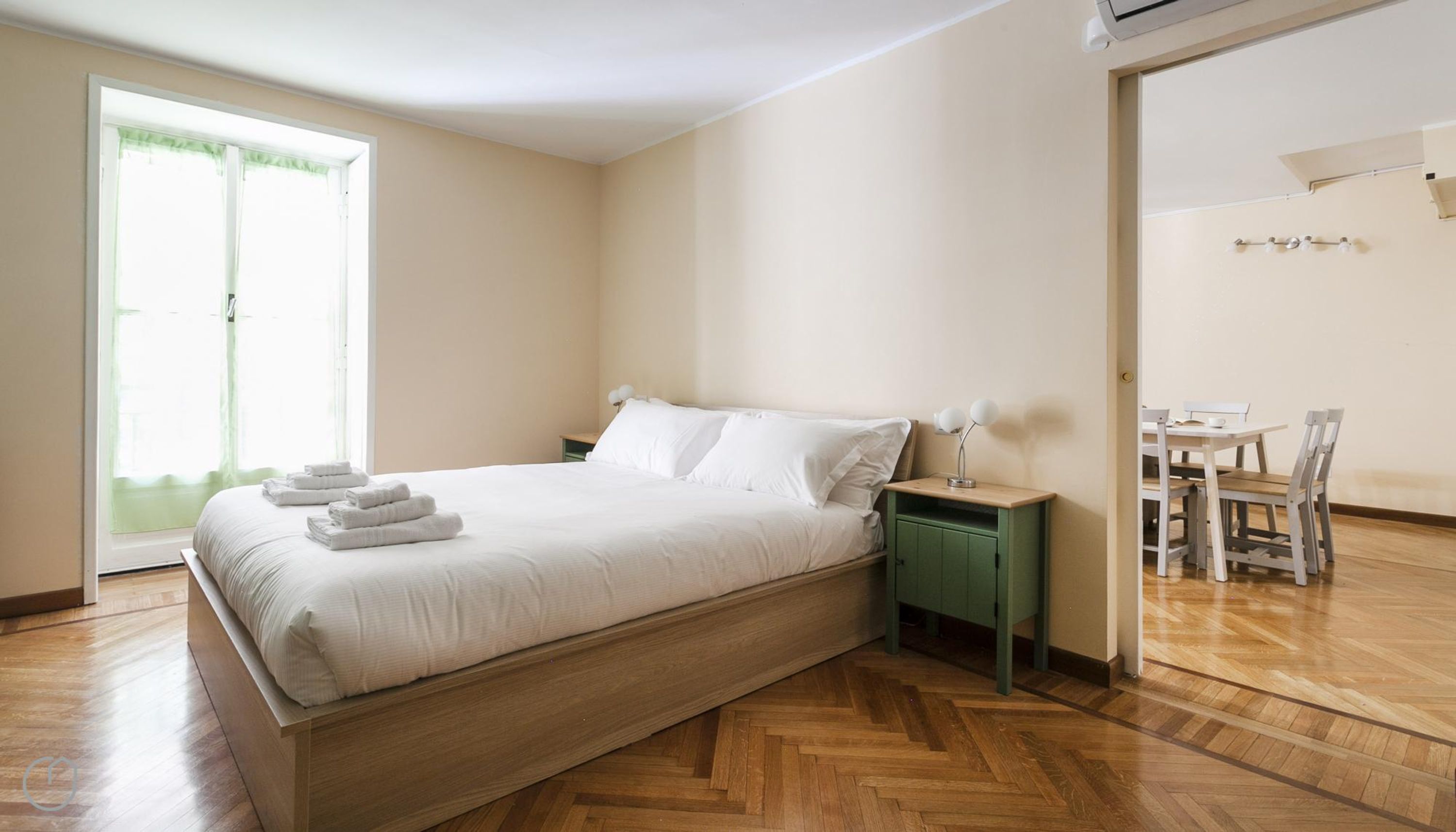 A bedroom with a wooden bed, a green nightstand, a bedside table with lamps, a wooden chair, and a small table with a chair.