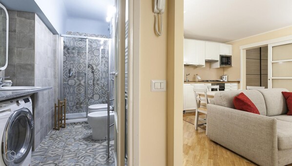 Un appartement compact doté d’une salle de bain avec douche, toilettes et lave-linge. La cuisine est équipée de meubles blancs et d’un four à micro-ondes. Un canapé beige agrémenté de coussins rouges se trouve dans l’espace de vie.