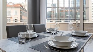 Een eettafel gedekt met borden, kommen, glazen en bestek in een modern interieur met grote ramen en uitzicht op gebouwen.
