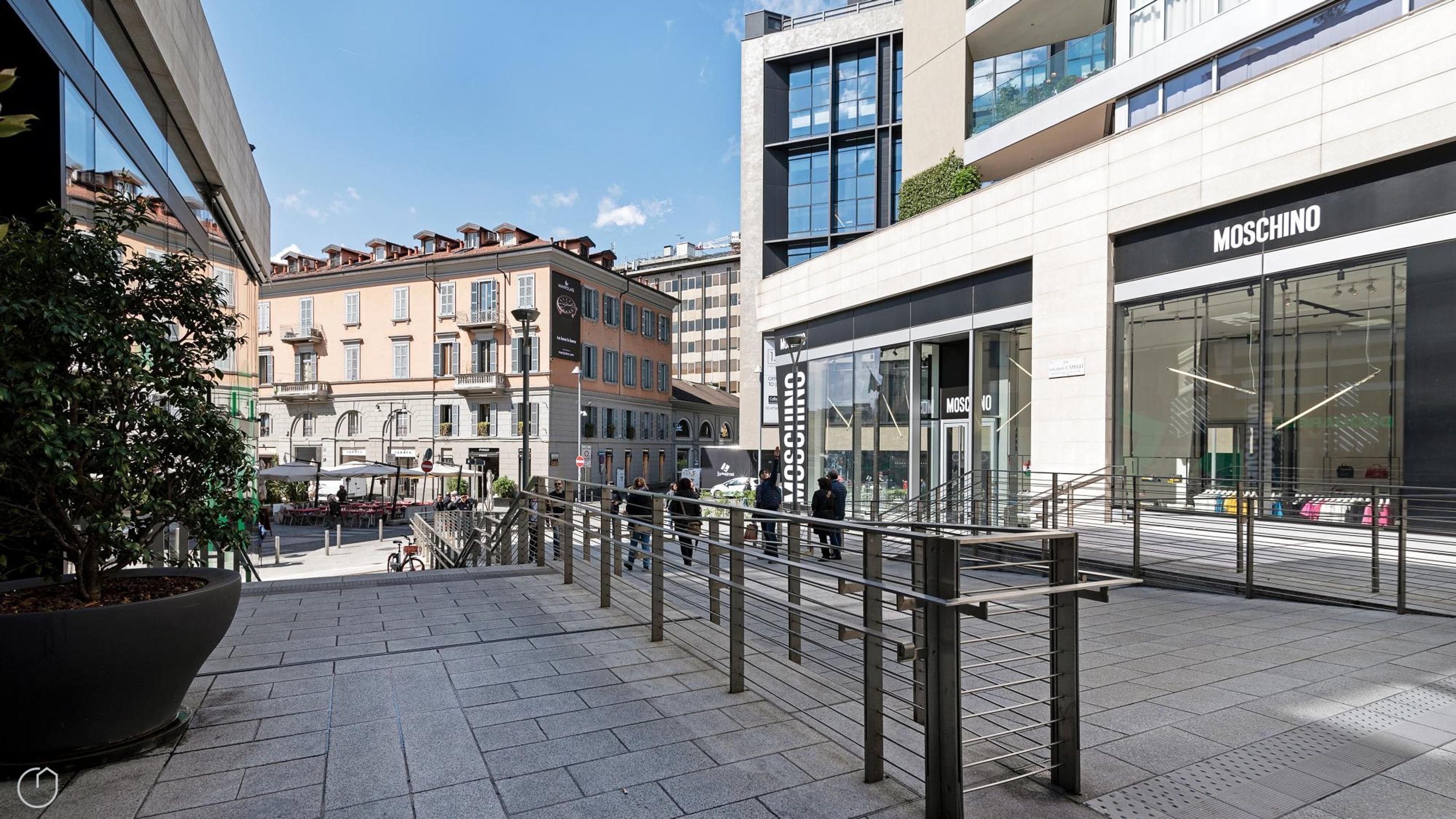 A modern urban street with a Moschino store, a large potted plant, and a pedestrian area with metal railings.