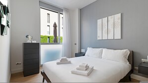 Een moderne slaapkamer met een groot bed, een dressoir, een nachtkastje en uitzicht op een gebouw door een raam.