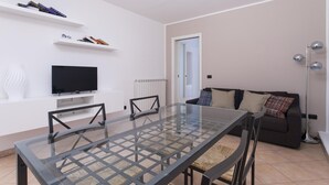 Een moderne woonkamer met een glazen eettafel, een flatscreen televisie, een bank en een vloerlamp.