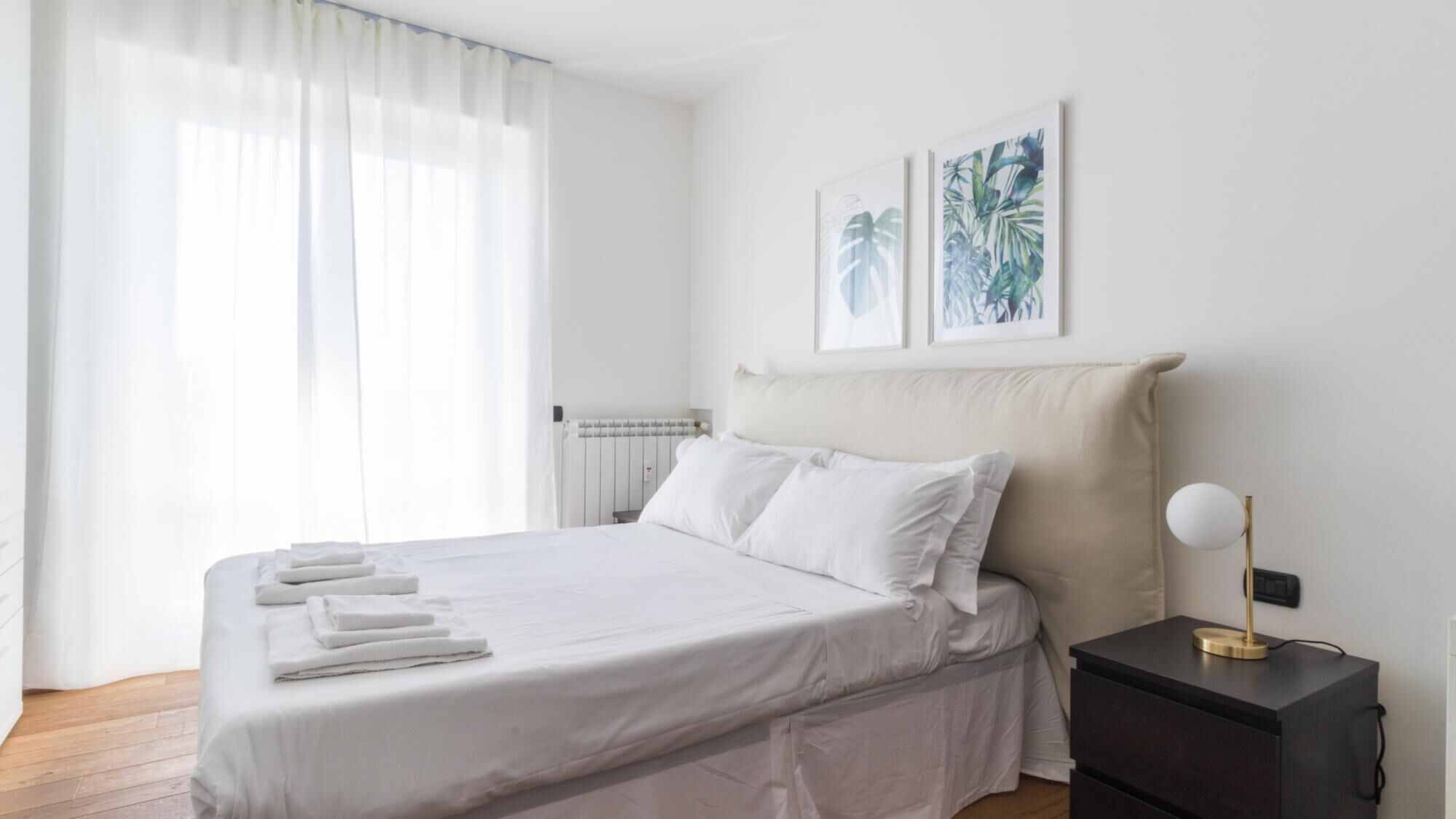 Una cama bien hecha con ropa de cama y toallas blancas, una mesita de noche con lámpara, y dos cuadros enmarcados en la pared.