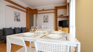 A dining area with a white table set for four, surrounded by white chairs. There are two paintings on the walls, a sofa with cushions, and a TV in the background. - Italianway Easy - Dei Tulipani 4 (Milan)