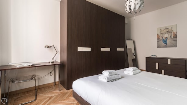 A modern bedroom with a wooden wardrobe, a desk with a lamp, a bed with neatly folded towels, and a painting on the wall. - Italianway Easy - Piceno 23 A (Milan)