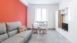 En moderne stue med en grå sofa, et lille spisebord med stole, et fjernsyn på et hvidt møbel og et stort vindue med hvide gardiner.