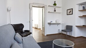 Un salon moderne avec un canapé gris, une table basse en verre et un fauteuil gris. On y trouve des étagères blanches ornées d’objets décoratifs et un tableau encadré accroché au mur.
