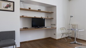 Une pièce avec un téléviseur à écran plat posé sur une étagère en bois, un fauteuil gris, une petite table avec une lampe et un radiateur.