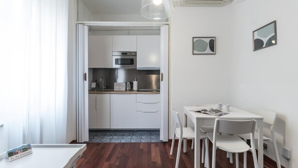 A compact kitchen with white cabinets, a microwave, and a coffee maker. There is a dining table with four chairs and a small table with a book and a cup.