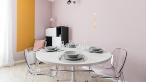 A dining area with a round table set for four, clear chairs, a white cabinet, and a pink wall. - Italianway Comfort - Buenos Aires 65 A (Milan)