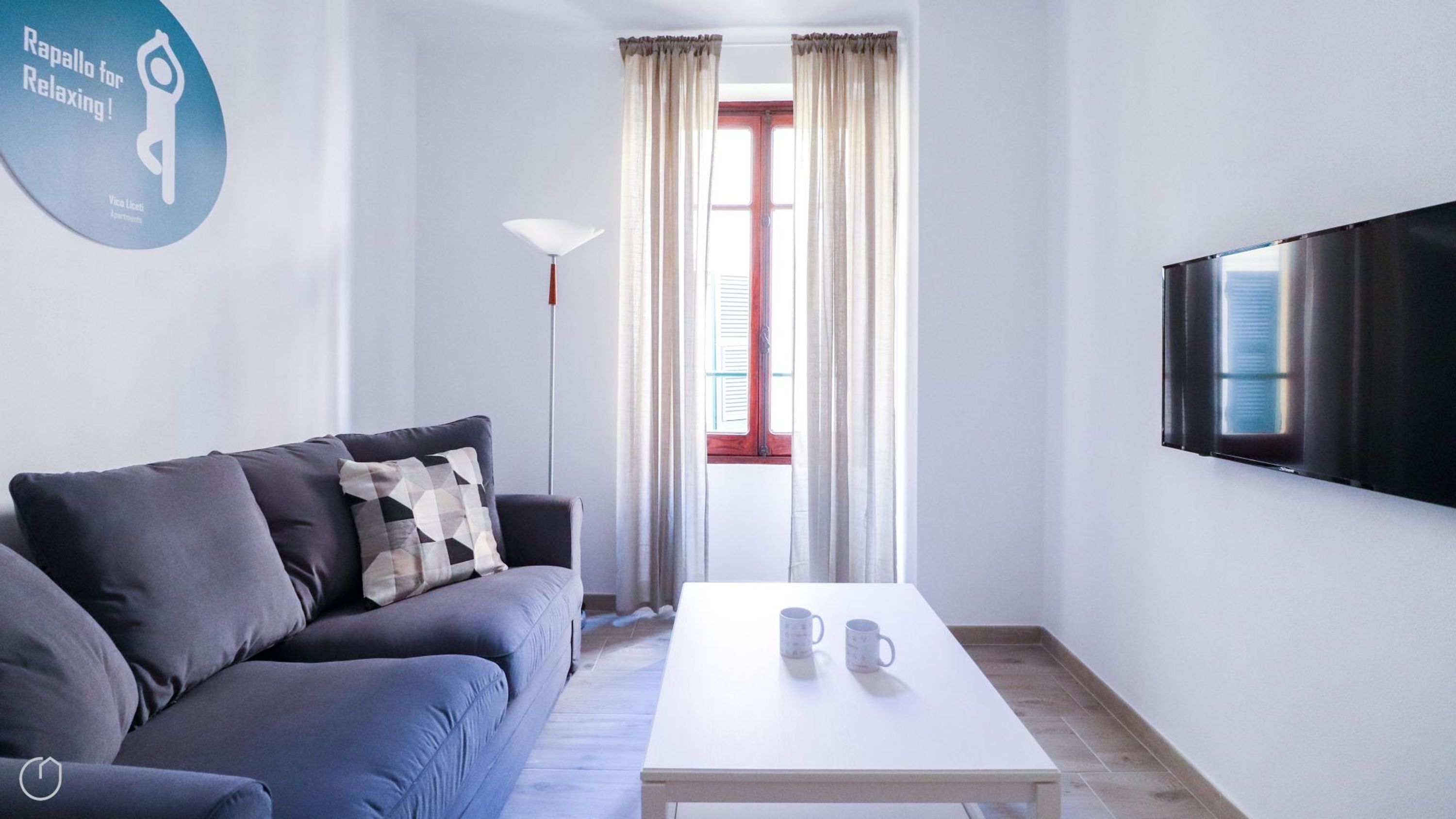 A modern living room with a sofa, a coffee table with two mugs, a floor lamp, and a TV mounted on the wall.