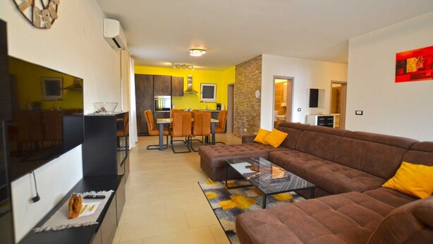A modern living room with a brown sofa, a glass coffee table, and a TV on a black stand. There is a kitchen in the background with yellow walls and a dining area.
