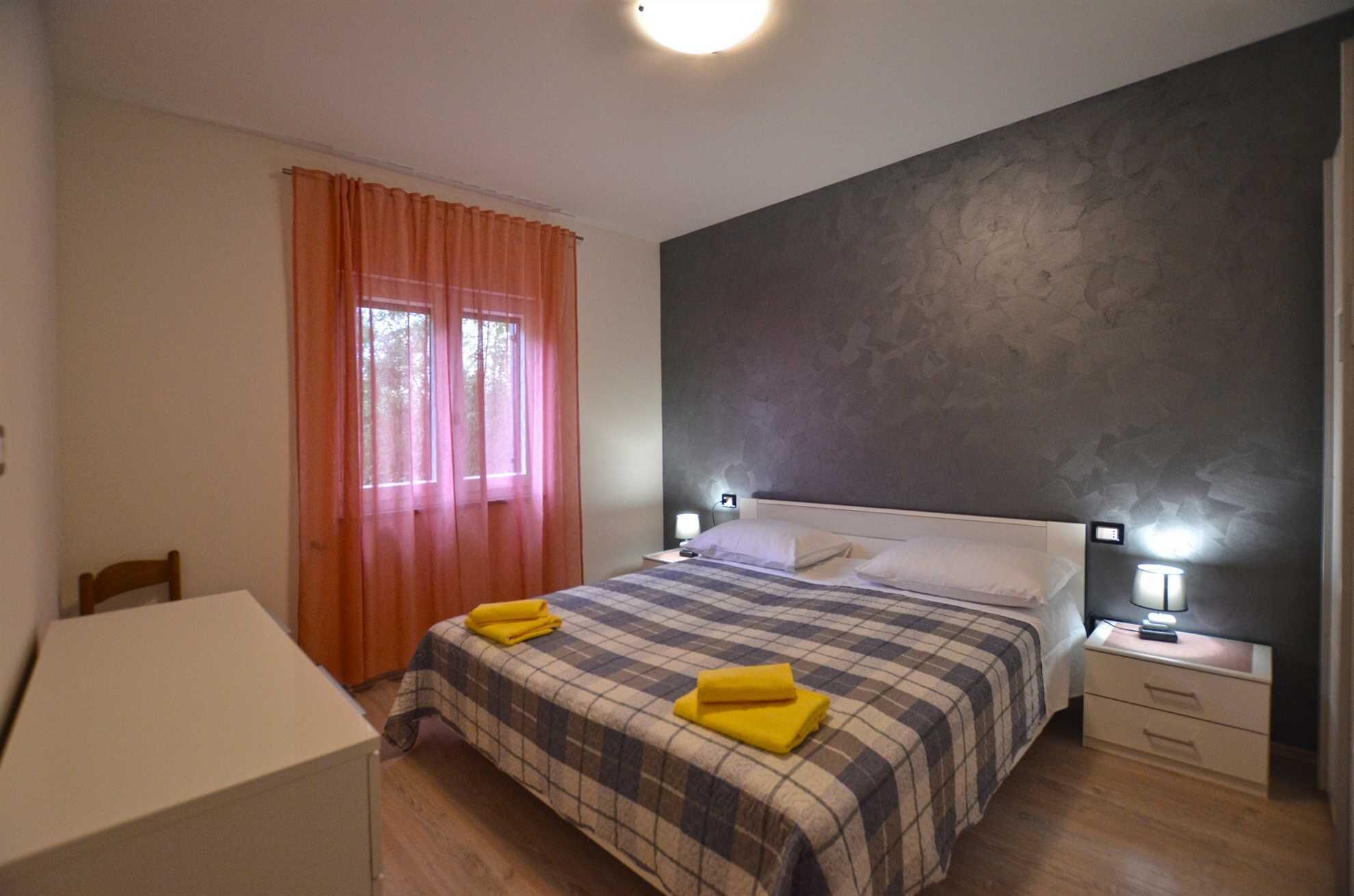 Un dormitorio con una cama, dos mesitas de noche con lámparas, una silla, un escritorio y una ventana con cortinas.