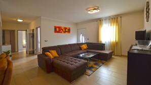 A living room with a sectional sofa, a coffee table, and a TV on a stand. There is a door leading to another room and a window with curtains.