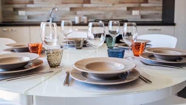 Une table de salle à manger ronde, mise avec des assiettes, des verres et des couverts, avec une cuisine moderne en arrière-plan.