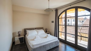 Une chambre à coucher comprenant un lit, des tables de chevet, une lampe et une grande fenêtre en arc d’où l’on peut admirer les bâtiments environnants.