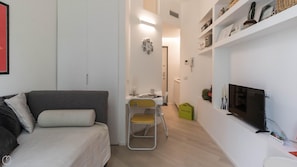 Un espacio de sala de estar pequeño y moderno, con un sofá, una silla plegable, un televisor y una estantería con diversos objetos.