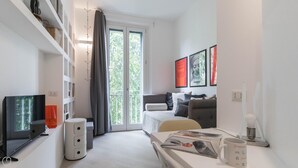 A modern living room with a sofa, coffee table, and a TV. There is a large window with curtains, a bookshelf, and a coffee machine. - Italianway Easy - Raffaello Sanzio 17 (Milan)