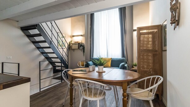 A dining area with a wooden table and white chairs, a staircase, a sofa with cushions, and a wall-mounted shelf with plants. - Chavez 2 (Milan)