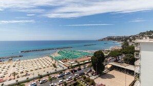 Vista de la playa con numerosos sombrillas y tumbonas, un aparcamiento y el mar con edificios a lo lejos.
