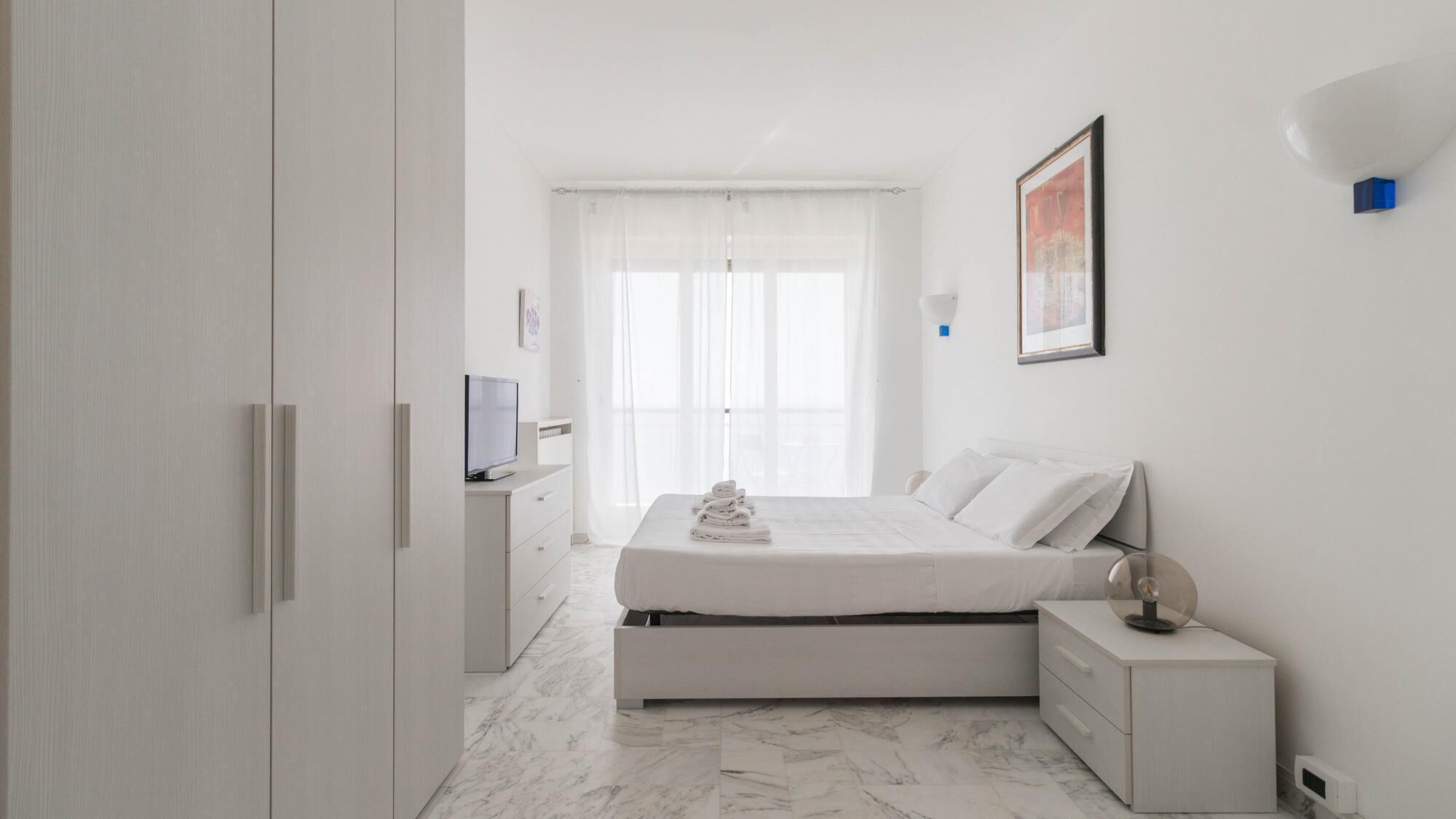Quarto moderno com cama, mesas de cabeceira, televisão e janela com cortinas leves.