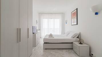 Quarto moderno com cama, mesas de cabeceira, televisão e janela com cortinas leves.