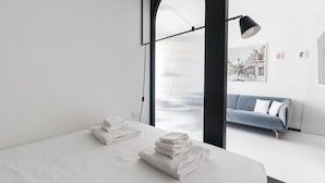 Een moderne hotelkamer met een bed, een stapel witte handdoeken, een blauwe bank en een groot raam met uitzicht op een gebouw.