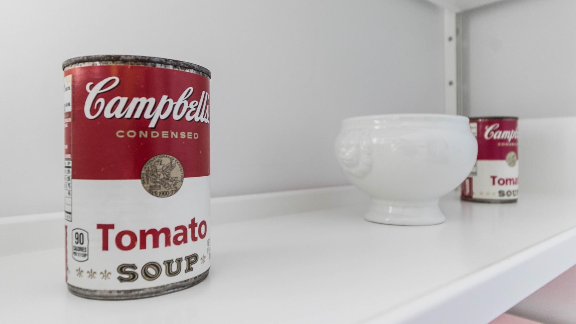 Uma lata de sopa de tomate Campbell numa prateleira, com uma chávena de cerâmica branca ao lado.