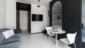 Een moderne woonkamer met een grijze bank, witte stoelen, een klein rond tafeltje en een aan de muur bevestigde televisie.