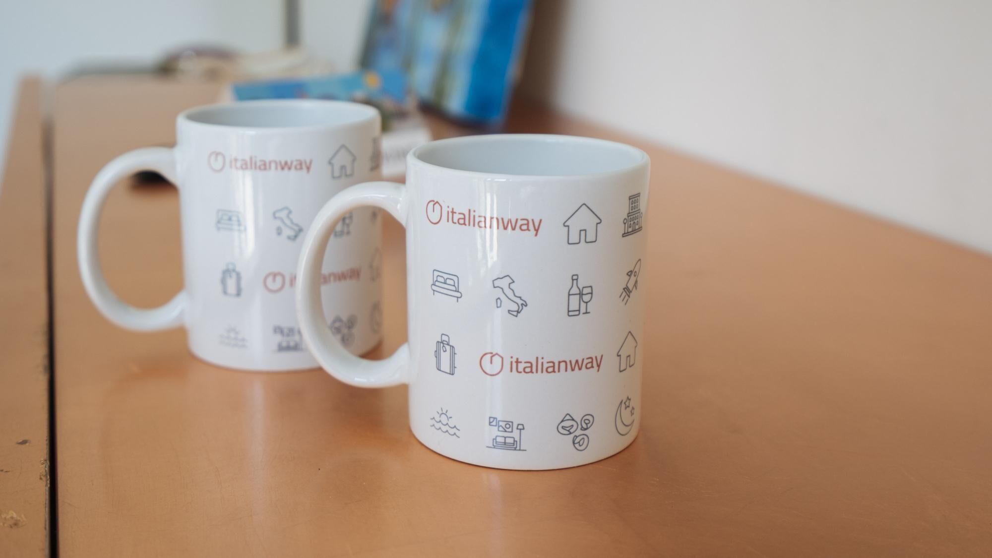 Deux tasses blanches ornées des logos d’Italianway et d’icônes liées au voyage, posées sur une surface en bois.