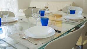 A dining table set with white plates, blue glasses, and cutlery, with a glass partition in the foreground. - Parco Villa Fiske 2 (Alassio)