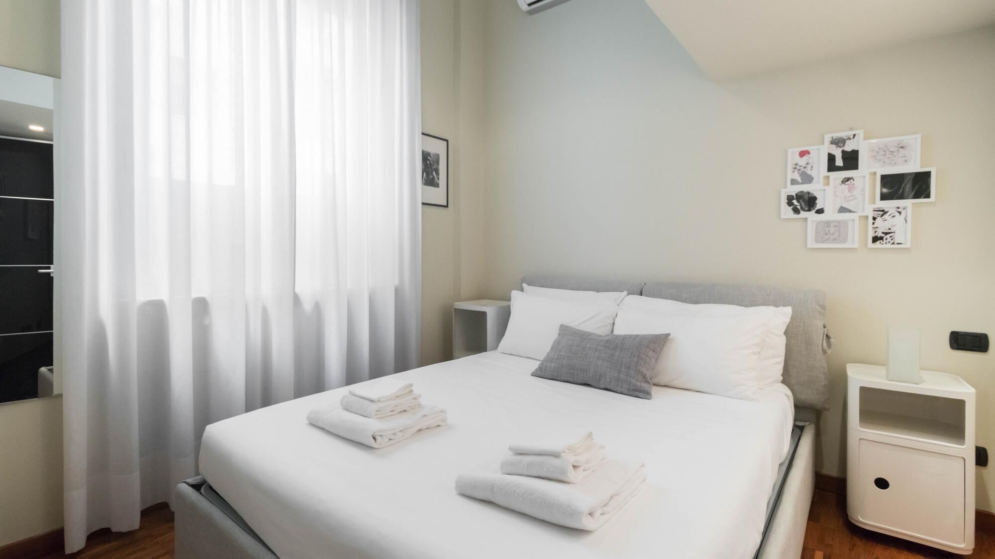 A neatly made bed with white linens and a grey pillow, a bedside table with a lamp, and a wall with framed pictures.