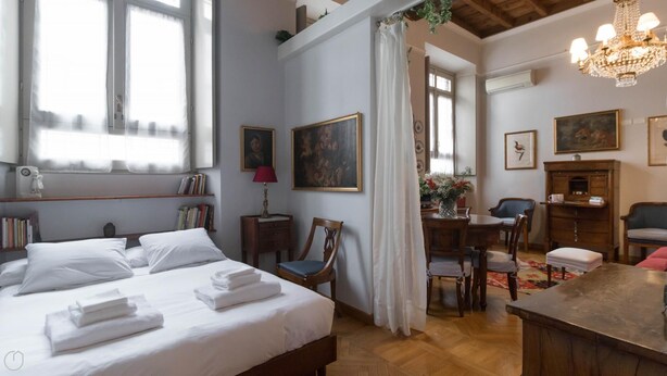 Un dormitorio con cama, mesita de noche y escritorio con sillas. Hay cuadros en las paredes y una araña.