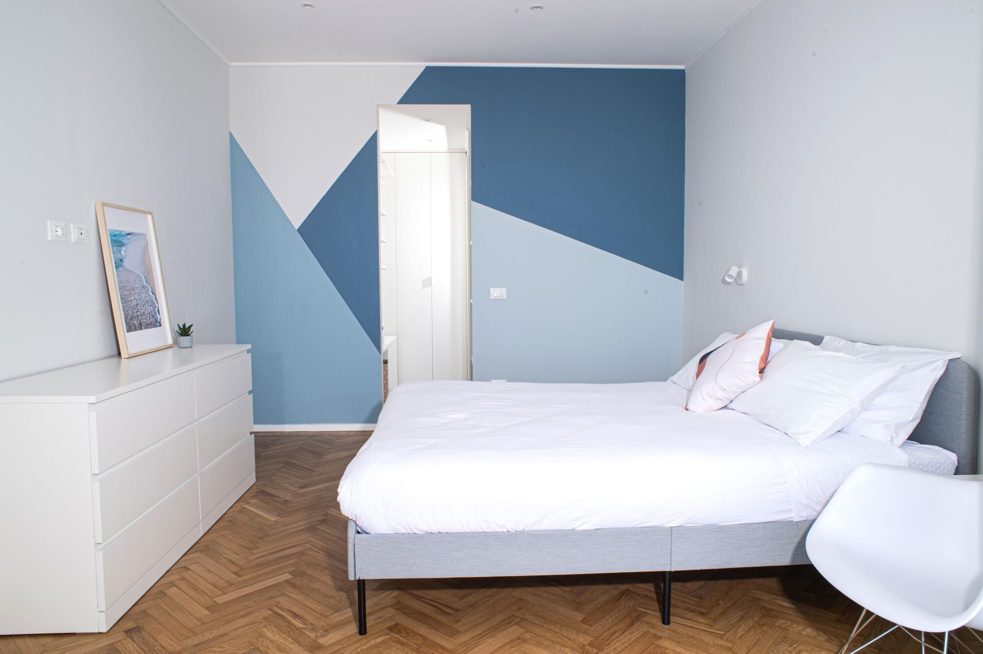 Un dormitorio moderno con una cama, un aparador blanco, una silla y un diseño geométrico en la pared.