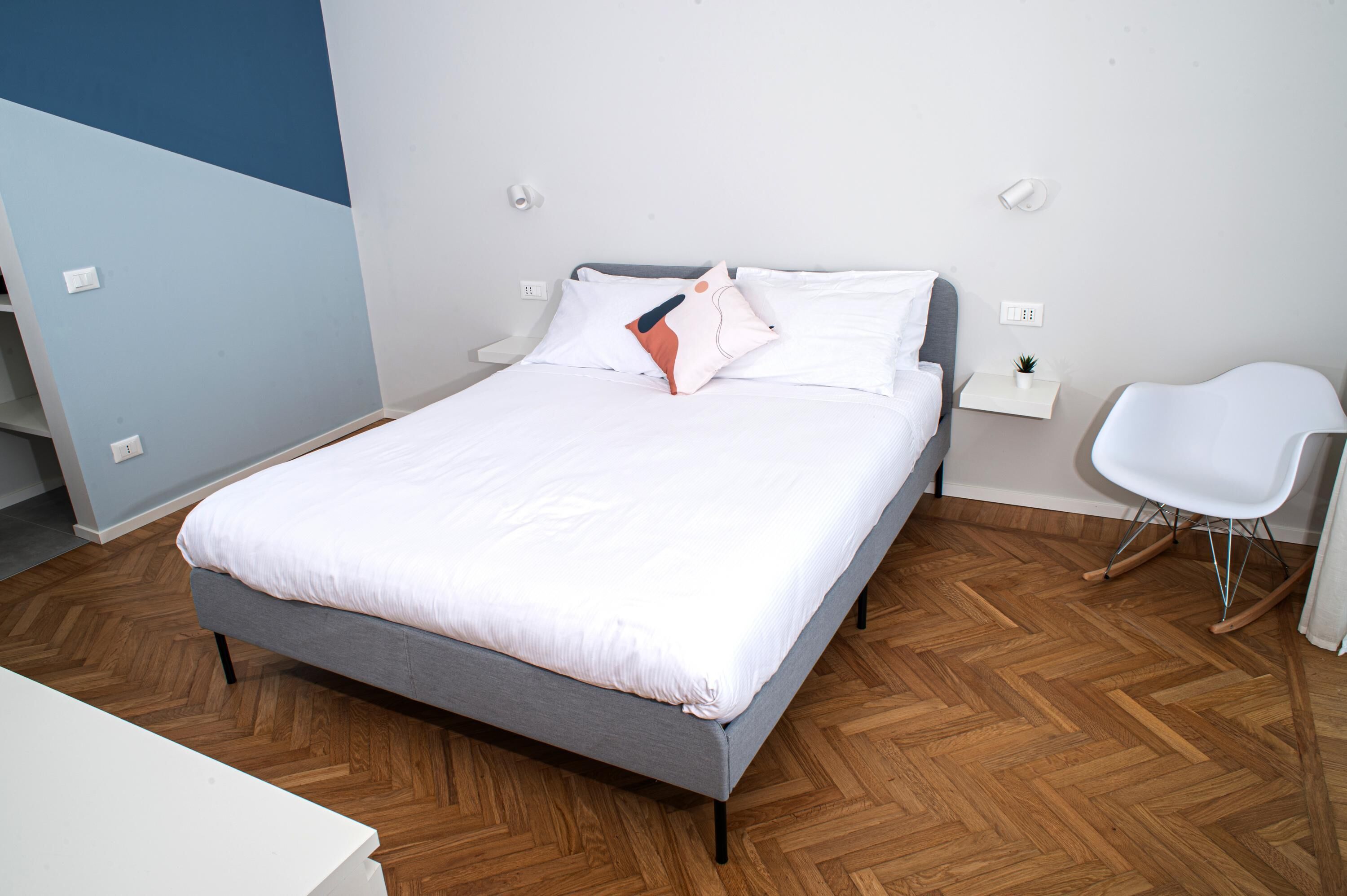 Quarto moderno com cama, mesas de cabeceira, uma cadeira e uma pequena planta.