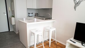 A compact kitchen with a white countertop, two stools, a kettle, and a TV on a stand.