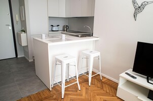 A compact kitchen with a white countertop, two stools, a kettle, and a TV on a stand.
