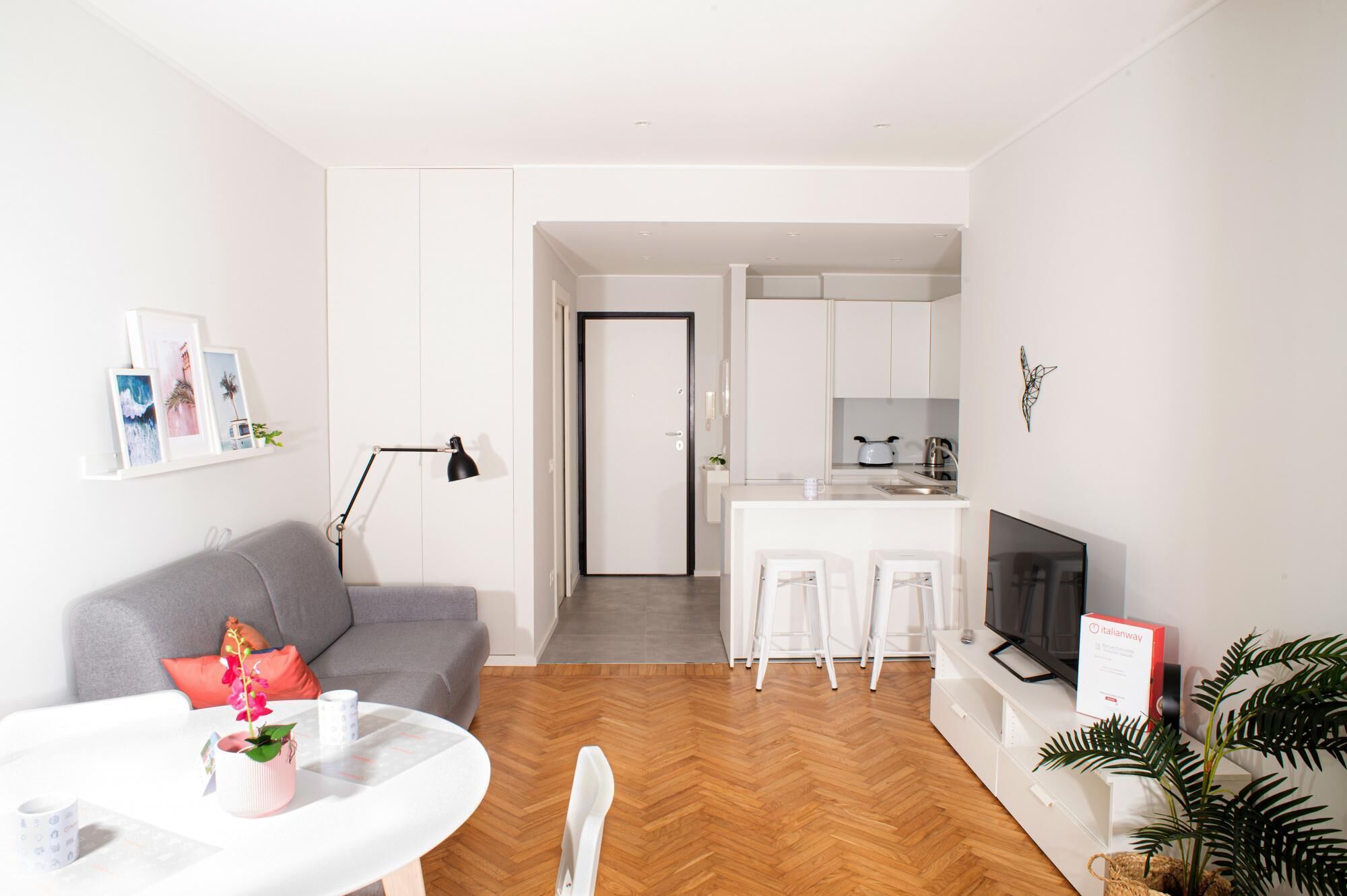 Un appartement moderne comprenant un salon, une cuisine et un coin repas. On y trouve un canapé gris, une table basse blanche et un téléviseur. La cuisine est équipée de meubles blancs et d’un plan de travail.