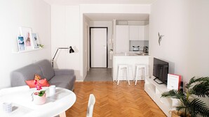A modern apartment with a living room, kitchen, and dining area. There is a grey sofa, a white coffee table, and a TV. The kitchen has white cabinets and a countertop.